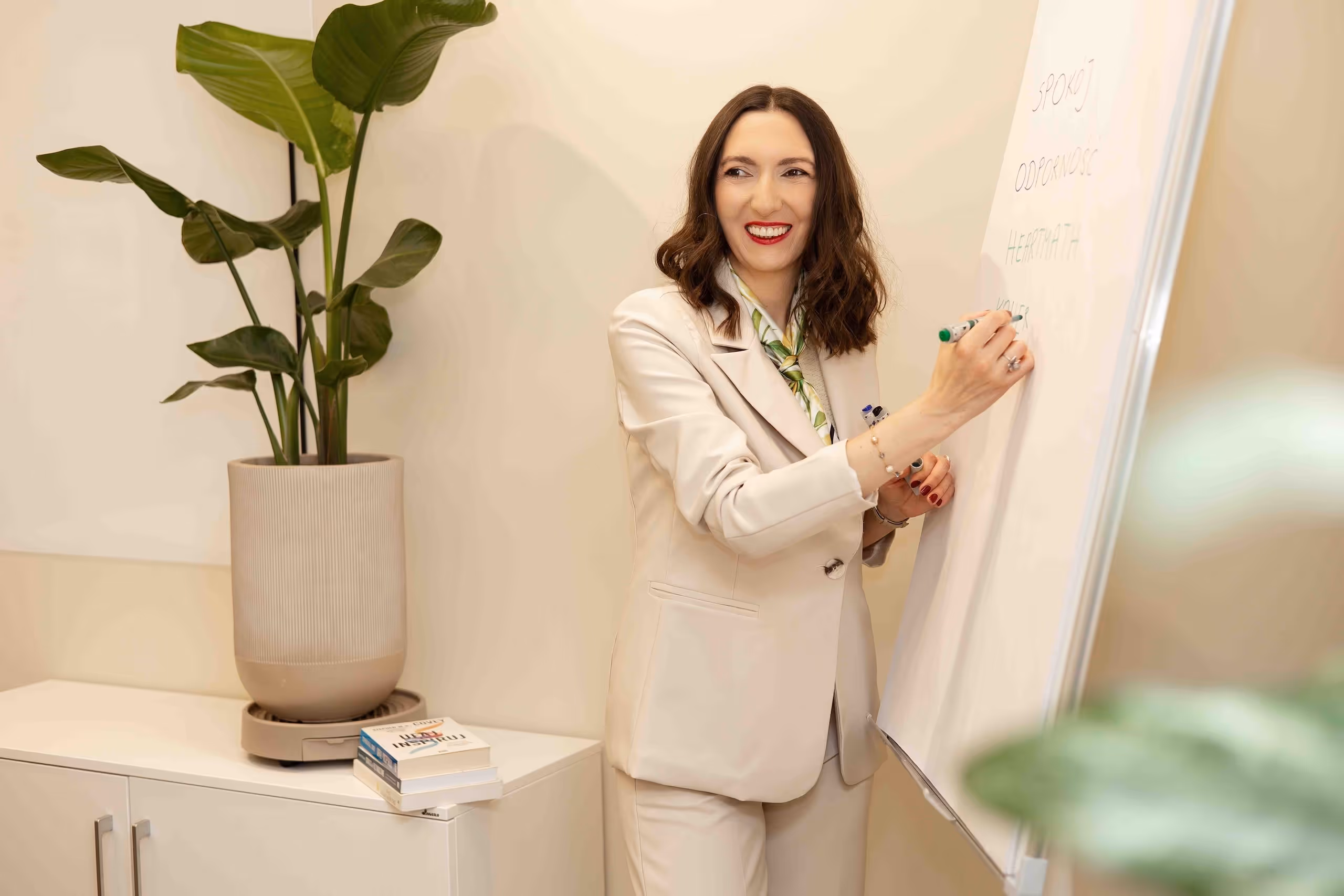 Alicja Dworowska w garniturze pisząca markerem na tablicy w jasnym pokoju z rośliną i książkami.