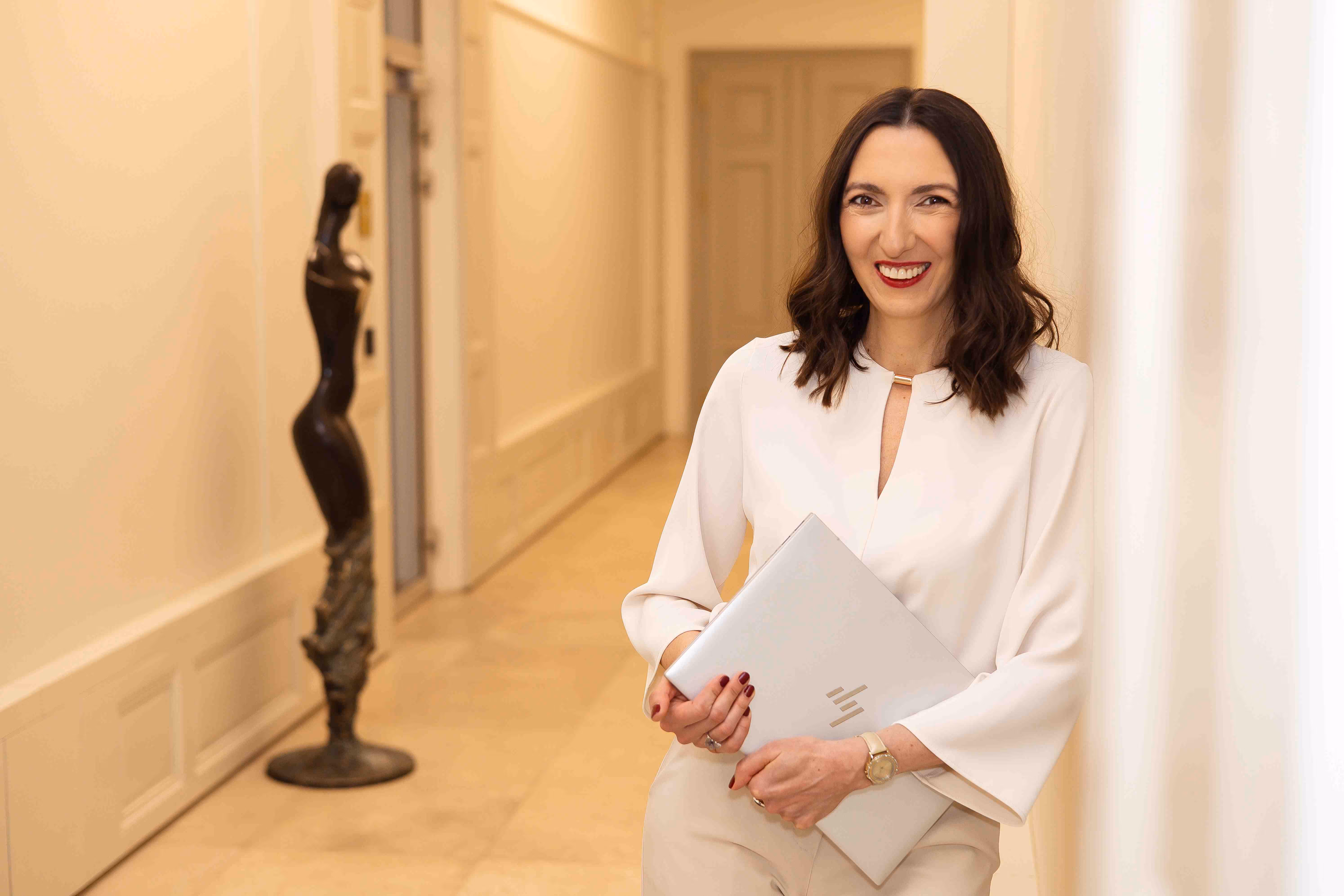 Alicja Dworowska w białej bluzce trzyma laptopa i uśmiecha się stojąc w jasnym korytarzu.